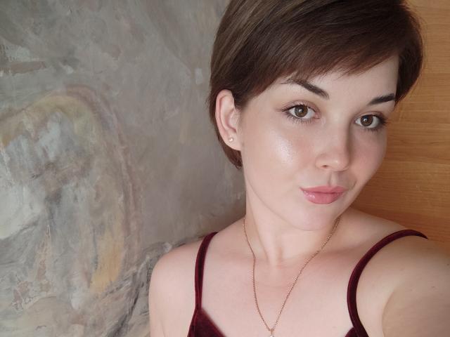 SweetyKitty69 has short brown hair, brown eyes, and wears a maroon top with a gold necklace. She's smiling with pink lipstick, showing a slight tan and cute earrings.