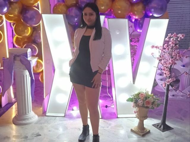 Karollteyes, com cabelo preto, vestindo uma jaqueta rosa, um mini-vestido preto e botas pretas até o tornozelo, está em frente a um sinal luminoso em forma de "V", com balões e flores..