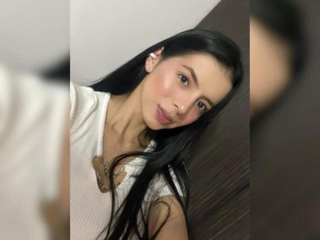 IsabellaJames has long black hair, a cute face, and is wearing a white shirt with a brown, decorative, butterfly-like necklace. She's leaning slightly to the right.