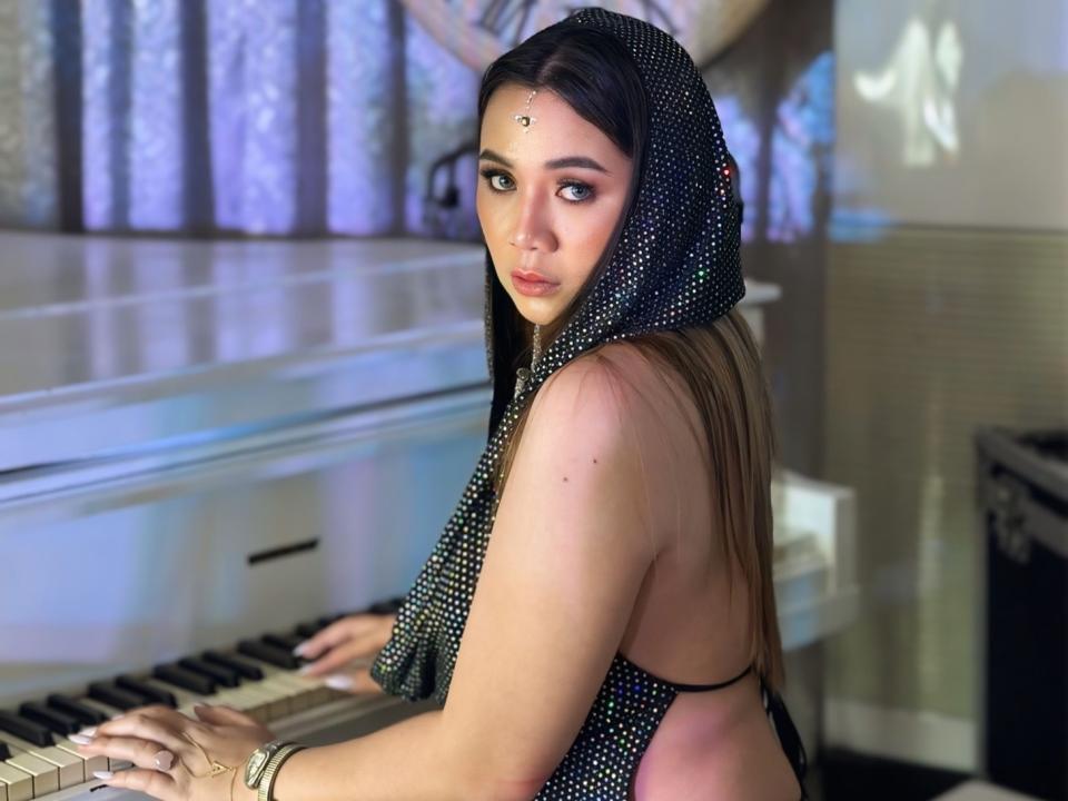 SabrinaMiler69 with dark hair, wearing a black polka dot hijab, playing a piano, has a nose ring, and a facial dot on her forehead, looking back with blue eyes.