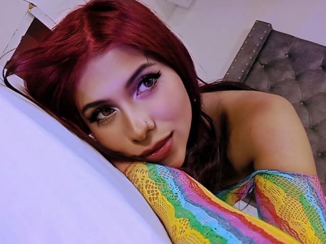 DanikaSerov with red hair, purple eyeliner, and nose ring, lying on a bed with colorful lace, looking up, her bare shoulders and pink lips visible.