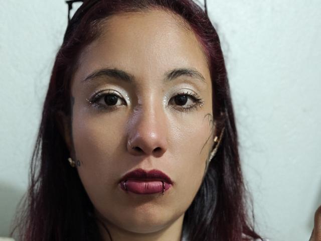 KataleyaNext's face with dark red lipstick, shiny white eyeshadow, and a nose piercing. Her dark hair is down, and she's wearing small earrings. Her expression is neutral.