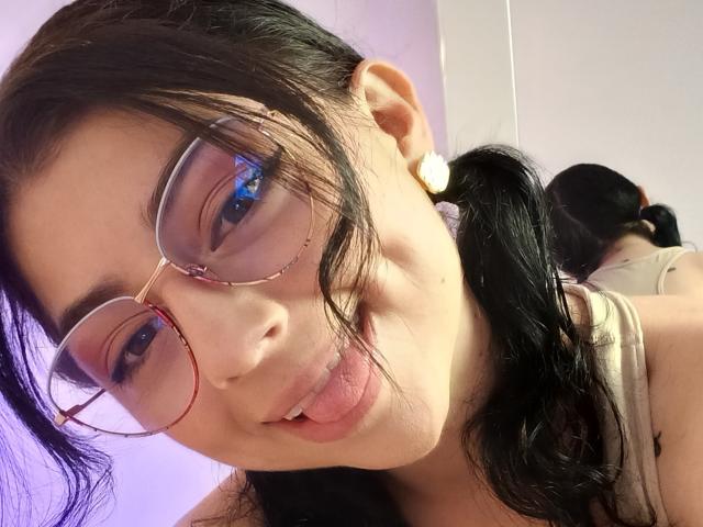 AnntonellaWatson with black pigtails, sticking out her tongue, wearing red-rimmed glasses and a beige top, showing her nose and lips. Her eyes are blue with pink eyeshadow.