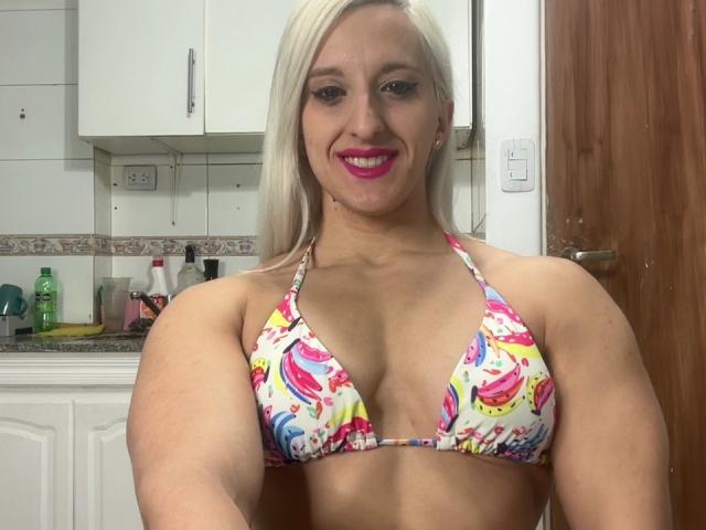 MuscleAngel with blonde hair, pink lipstick, and colorful tie-dye bikini top, showing off her large, firm tits and muscular arms in a kitchen.