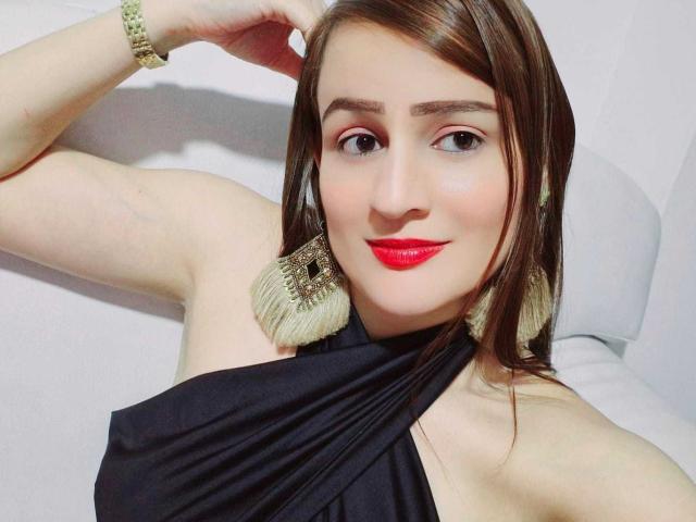 CamilleDurand with red lipstick, black halter top, and large tassel earrings, smiling, brown hair, arm raised, wearing a gold bracelet, fair skin, and red eyeshadow.