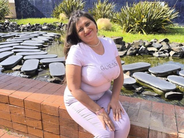 BlissSophia with large, saggy tits in a tight, pink shirt and matching pants, sitting on a brick wall by a stone-paved water feature, smiling.
