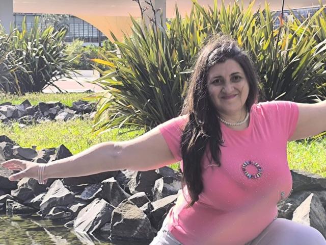 BlissSophia has big boobs, dark brown hair, and a pink shirt with a flower emblem. She's kneeling by rocks, arms out, smiling, wearing a pearl necklace.