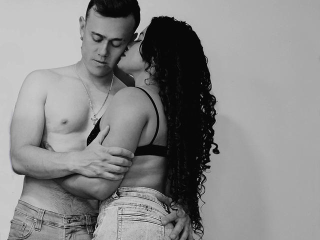 JenAndPatrick with curly black hair, kissing Patrick's neck, while he holds her against his chest, both shirtless, Jen in a black bra, Patrick in jeans, necklace on Patrick.