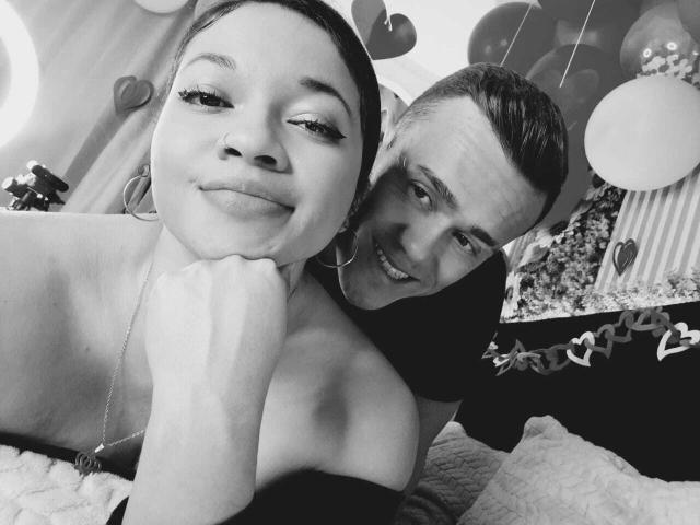 Black-and-white selfie of JenAndPatrick, topless with puffy tits, wearing hoop earrings, Patrick with slicked-back hair, smiling, leaning over her shoulder. Balloons and hearts in background.