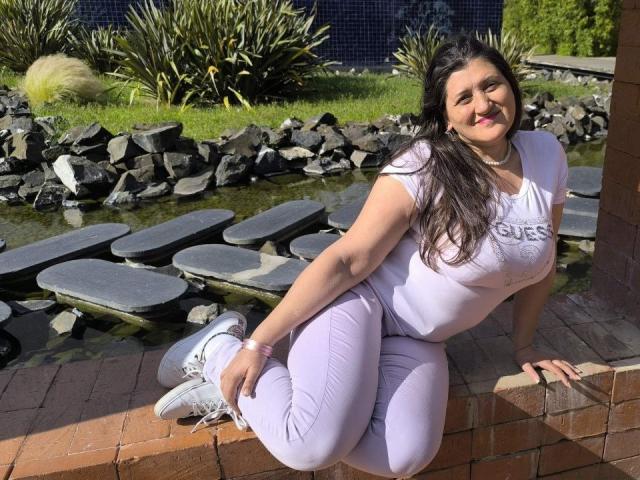 BlissSophia, with big tits and ass, kneels in sunlight, wearing white shirt and pants, long dark hair, smiling, by a stone water feature, white sneakers, brick ledge.