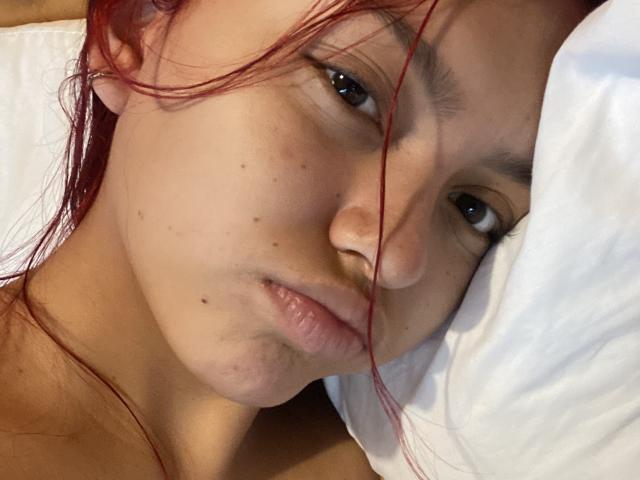 AlyMartiny with red hair, lying on a white pillow, has freckles, small moles, and a pouty face, showing slightly parted lips and a neutral expression.