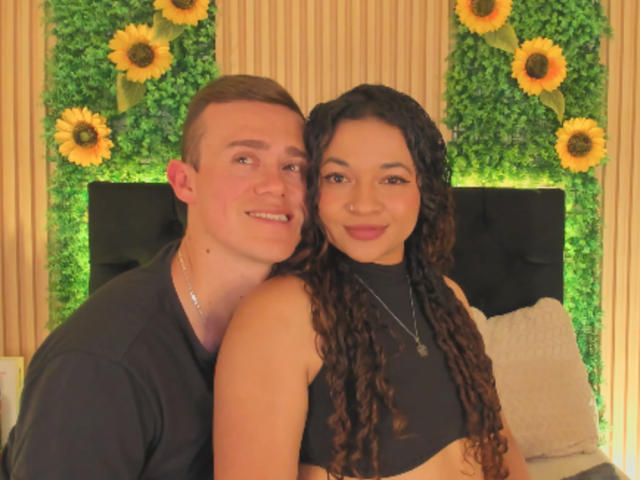 JenAndPatrick with curly brown hair and dark top, and Patrick with short brown hair and black shirt, smiling, leaning on a black headboard with sunflowers and greenery.
