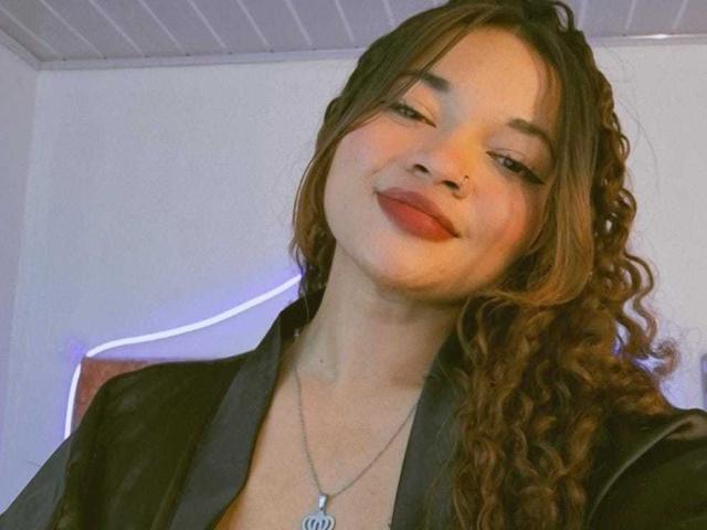 JenAndPatrick with curly brown hair, nose ring, and red lipstick, wearing a black leather jacket, smiling slightly, and a necklace with a pendant, in a selfie.