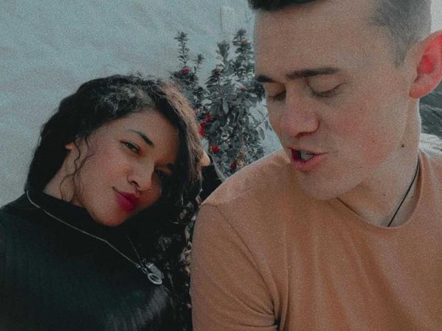JenAndPatrick, with curly black hair and red lipstick, leans on a pale guy in a beige shirt, both looking down. She's got a black jacket, he's wearing a necklace.