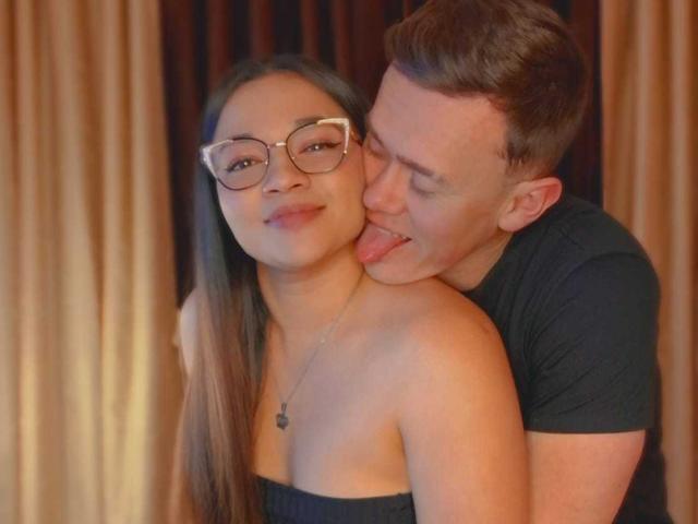 JenAndPatrick with glasses, long brown hair, black strapless top, and Patrick with short brown hair, black t-shirt, tongue out, hugging her from behind, showing affection.
