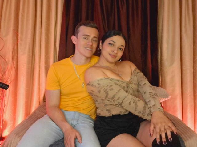 JenAndPatrick, a cute couple, sitting together. Jen has big boobs, black hair, and a beige off-shoulder top. Patrick, with short brown hair, wears a yellow tee and jeans.