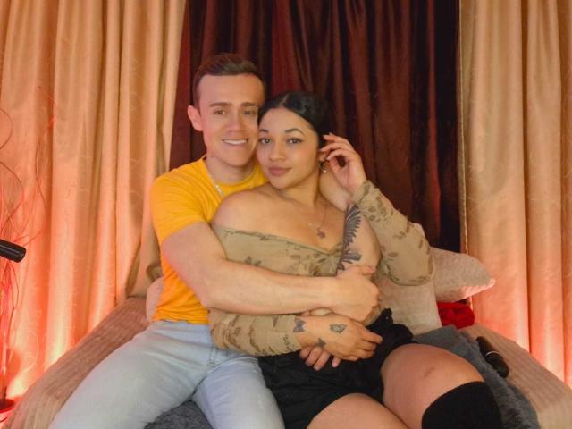 JenAndPatrick, with her tits slightly visible, sits on a bed, arms around Patrick, who's wearing a yellow tee, in a cozy, intimate setting. He's behind her, smiling.