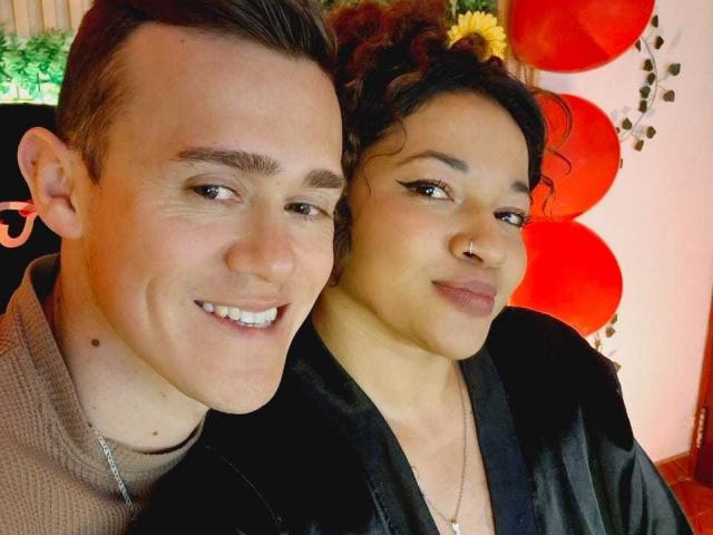 JenAndPatrick have cute faces, Jen's tits are covered, Patrick's ass is not, they're smiling, Jen's in a black robe, Patrick's in a brown sweater, red balloons in background.