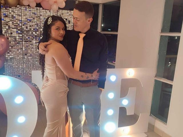 JenAndPatrick, a sexy woman with long black hair and tits in a satin pink dress, stands in an embrace with Patrick, a cute guy in a black shirt and yellow tie.