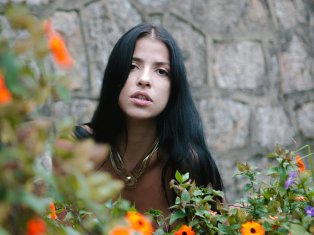 MistressHellvira with long black hair, brown top, and gold necklace, surrounded by orange flowers and green leaves, looking slightly up with a neutral expression.