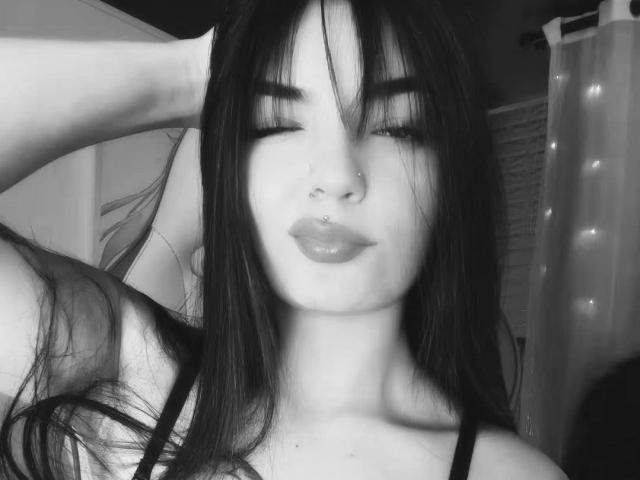 Black-and-white photo of MiaXSmithy with dark, straight hair, eyelashes down, nose ring, lip ring, glossy lips, hand in hair, wearing a black top, slightly visible cleavage.