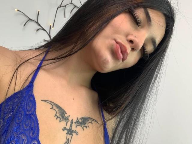 MiaXSmithy with long black hair, blue lace top, nose ring, and bat tattoo on chest, puckering lips. Her tits are small, and she has long eyelashes.