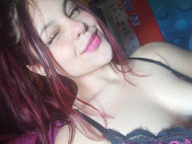 Sophie Hill, avec ses cheveux rouge-rose, son anneau de nez et son rouge à lèvres rose, clignant de l'œil, portant un haut en dentelle noire qui révèle ses seins, souriant doucement, peau claire, dans une pose détendue et décontractée..