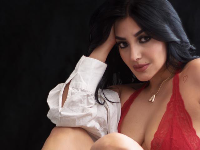 ArianaCortes with dark hair, wearing a red lace bra, white shirt, and gold necklace, looking at the camera, her hand on her head, her tits visible.