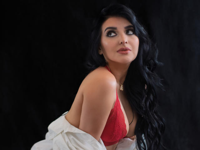 ArianaCortes with long black hair, wearing a red lace bra, white shirt off her shoulders, looking up with a slight smile, showing her tits, against a black background.