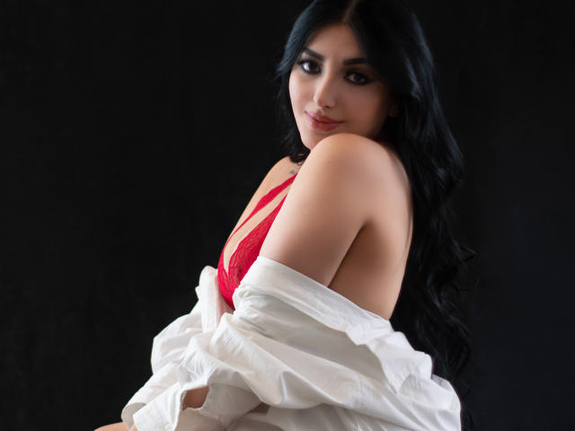 ArianaCortes with long black hair, red lace bra, and white shirt falling off her shoulders, looking back with a slight smile, showing her bare shoulder and cleavage.