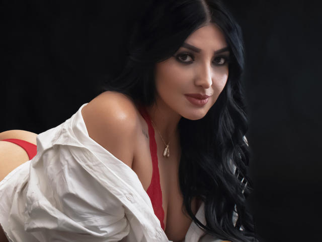 ArianaCortes with long black hair, red bra, and off-shoulder white shirt, looking seductively at the camera, showing her tits and a glimpse of her ass.