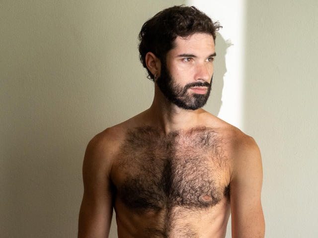 PupScritch è un uomo senza camicia, con il petto peloso, capelli castani ricci e barba, in piedi contro un muro chiaro, che guarda di lato con un'espressione seria..