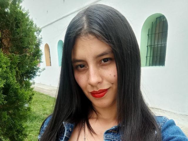 Madisson Fontaine, con i capelli neri lisci, rossetto rosso e una giacca di jeans, mostra una piccola lentiggine sulla guancia, ed è in piedi accanto a un cespuglio verde..