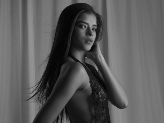 Jessica Lopes, com cabelos longos e escuros, vestindo uma blusa com amarração nas costas, vista lateral, mão perto do rosto, olhando para a câmera, preto e branco, luz suave, sexy, confiante..