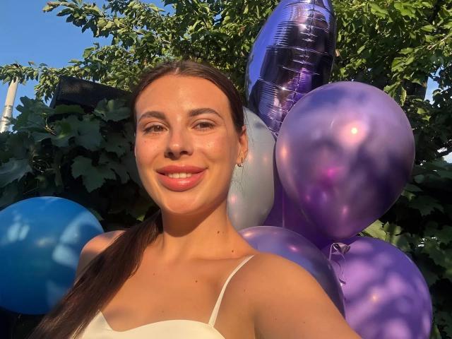 MoonriseGoddessx-hot with brown hair, smiling, wearing a white top, surrounded by purple and blue balloons, and a shiny purple balloon in the background. Sunlight highlights her face.