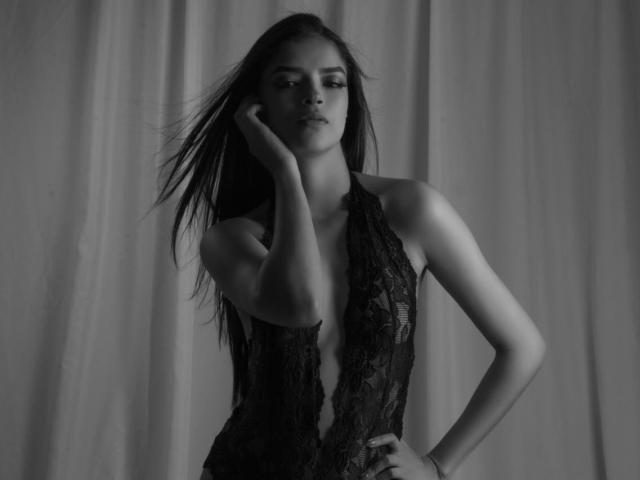 Jessica Lopes, con cabello largo y oscuro, vestida con un body negro de encaje, con una mano en la mejilla, mirando hacia abajo, de pie frente a una cortina, en blanco y negro, sus pechos visibles a través del encaje..