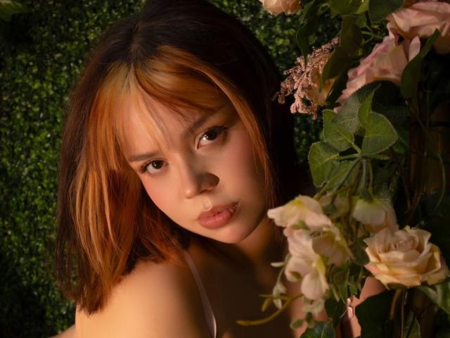 HanniRoss has brown and blonde hair, pouty lips, and is looking down with a serious expression. She's surrounded by pink roses and green leaves.