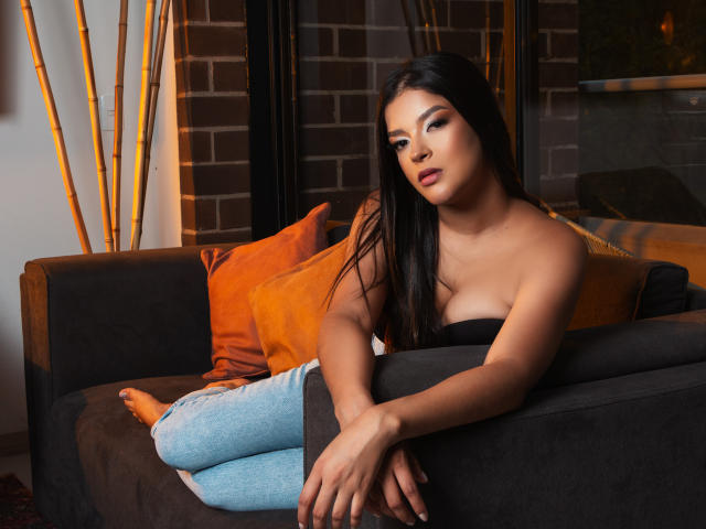 Jessica Lopes, com cabelos longos e pretos, pele bronzeada e seios grandes, vestindo uma blusa preta sem alças e jeans azul claro, sentada em um sofá marrom escuro com almofadas laranja..