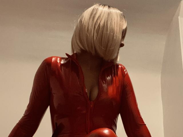 MilaDeesse with blonde bob, wearing shiny red latex top, zipped down, showing cleavage. Red latex reflects light, skin is dark. She's looking down, head tilted.
