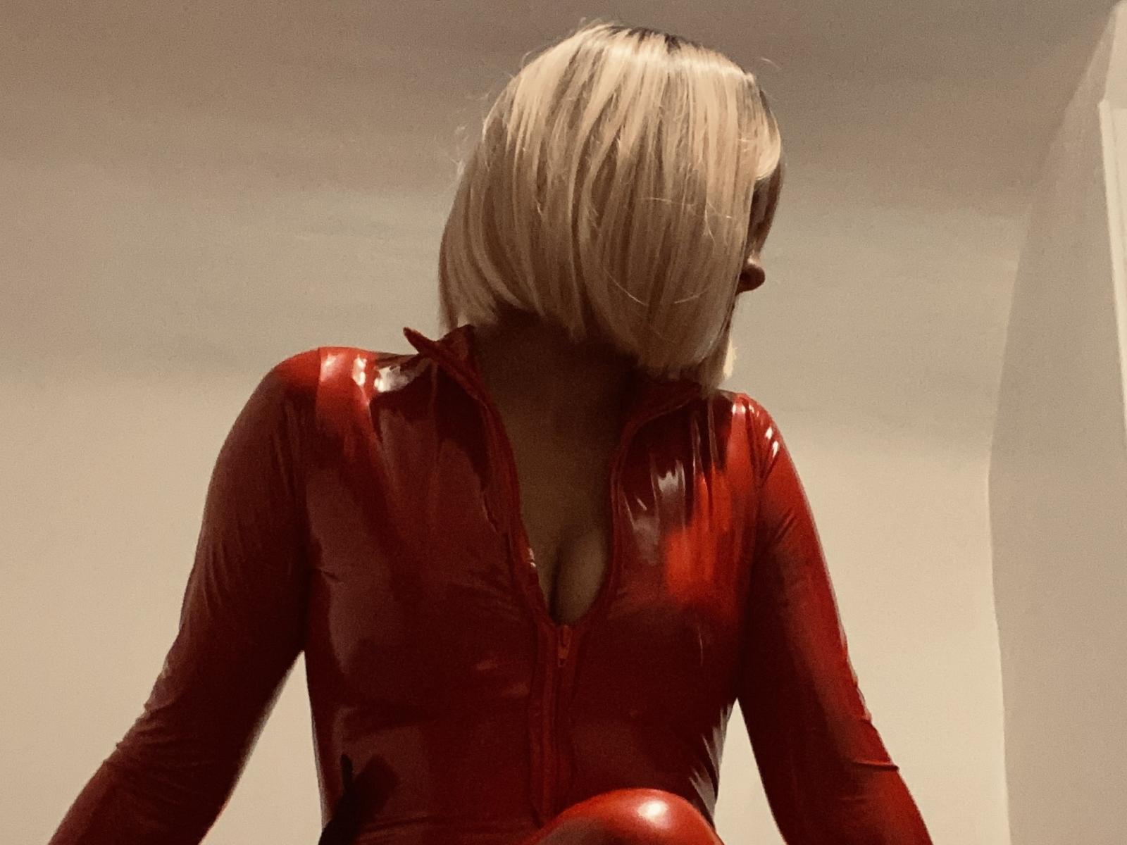MilaDeesse with blonde bob, wearing shiny red latex top, zipped down, showing cleavage. Red latex reflects light, skin is dark. She's looking down, head tilted.