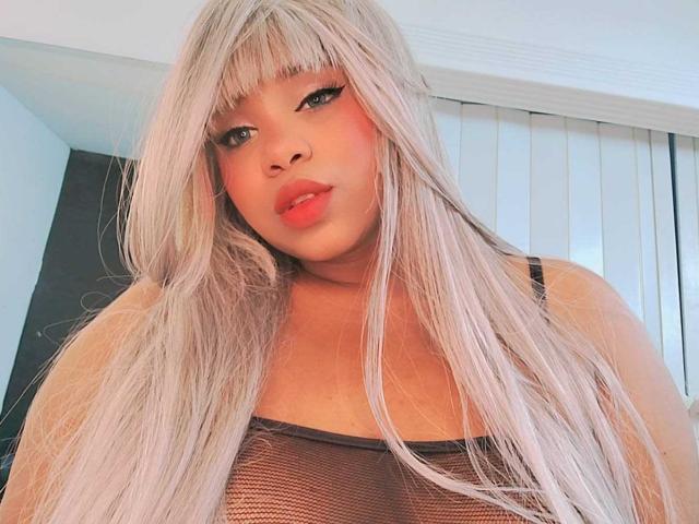 LizzyFoxy with long platinum blonde hair, wearing a black mesh top, showing her tits, has pink blush on her cheeks, and full pink lips, looking at the camera.