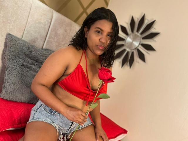 SahraCandy, with medium brown skin and curly black hair, wears a red halter top and frayed denim shorts, holding a red rose, sitting on a bed with red pillows.
