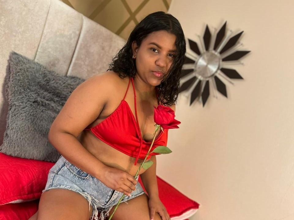 SahraCandy, with medium brown skin and curly black hair, wears a red halter top and frayed denim shorts, holding a red rose, sitting on a bed with red pillows.
