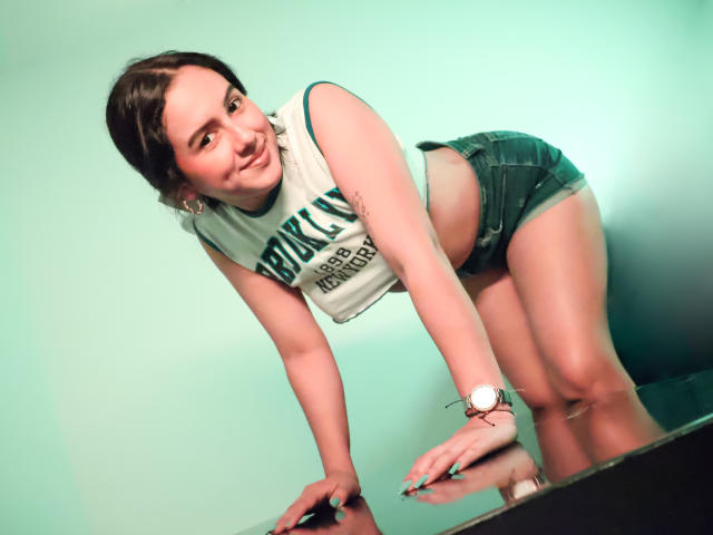 EmmieBellefort, vorgebeugt, trägt ein weißes Top ohne Ärmel und Jeansshorts, lächelt in die Kamera und hat eine Armbanduhr am linken Handgelenk, vor einem grünlichen Hintergrund..