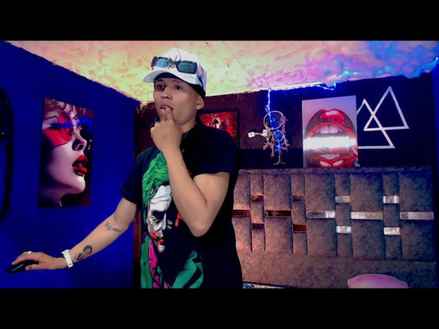 DanielTaylor, wearing a white cap and black shirt with green print, stands with finger to lips, sporting tattoos and a wristband, in a colorful, eclectic room.