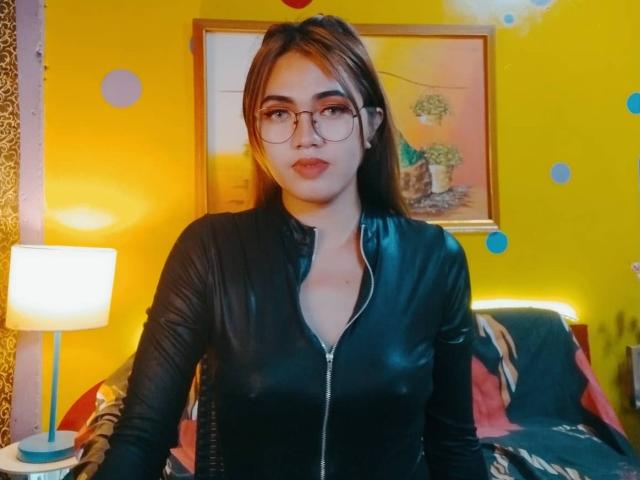 AsianHotCock69 with glasses, wearing a black leather zip-up, showing her tits, in a yellow room, standing in front of a bed with a lamp on the left.