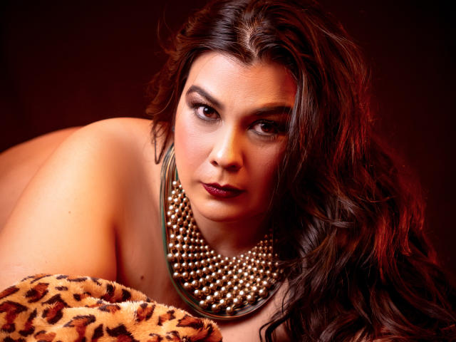 MartinaFox69 with long brown hair, dark lipstick, pearl necklace, lying on leopard print, looking seductively at camera. Boobs partially visible, brown eyes, serious expression.
