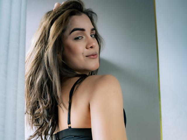 AliceCoolenk with wavy brown hair, black bra, and a slight smile, looking over her shoulder, her hand in her hair, showing her tits and a confident expression.