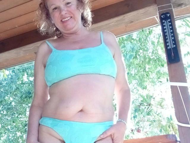 Jutta1-hot has pale skin, curly brown hair, and wears a light blue bra and panties set. Her small tits and slightly chubby belly are visible. She's smiling.