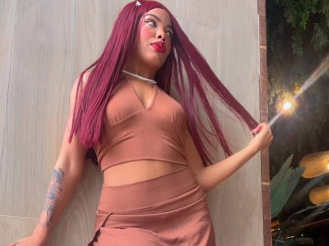 LexieBloom with long red hair, wearing a brown crop top and matching skirt, holding her hair, showing a tattoo on her left arm, and red lipstick.